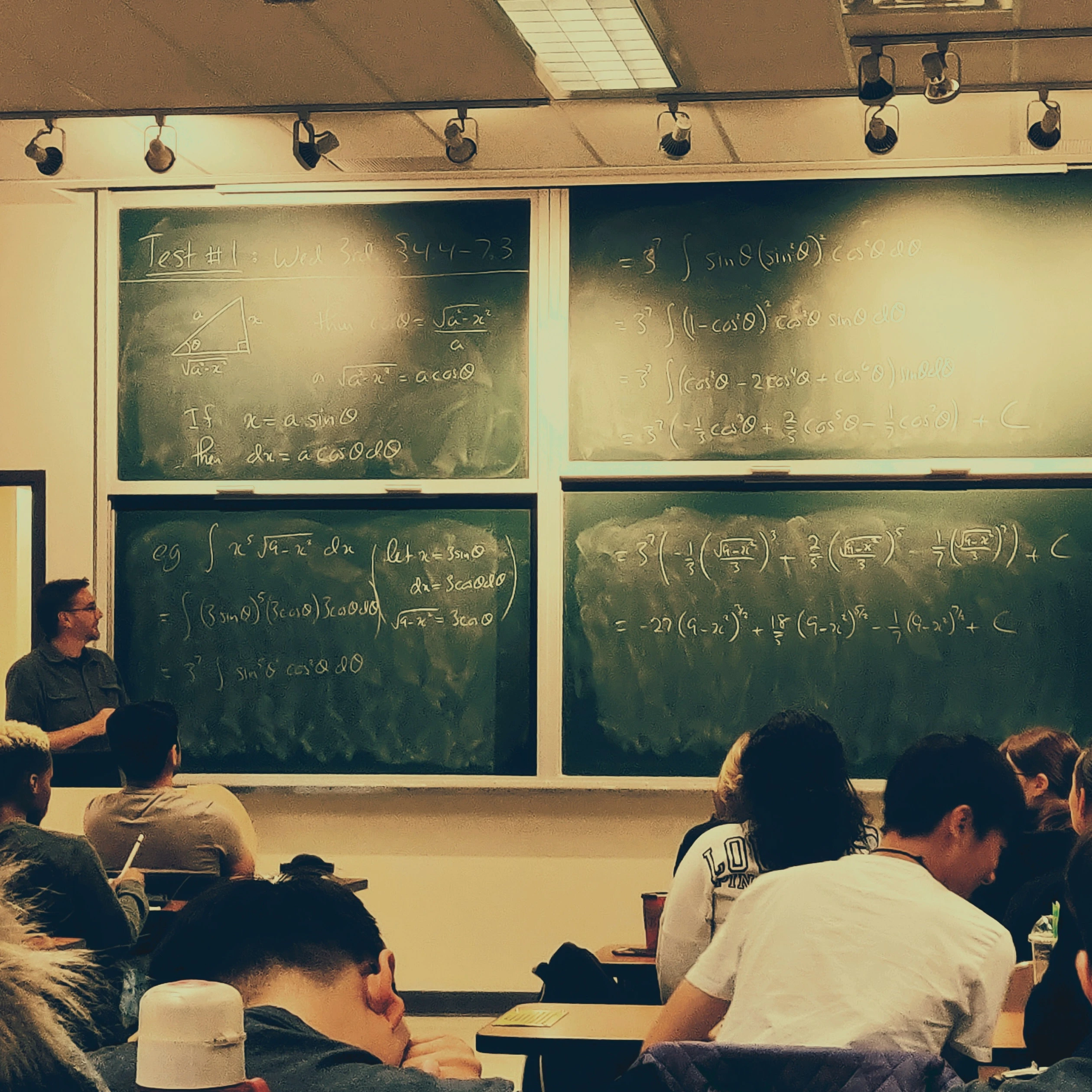 Imagem de sala de aula com alguns alunos focados e um professor explicando matéria na louza.